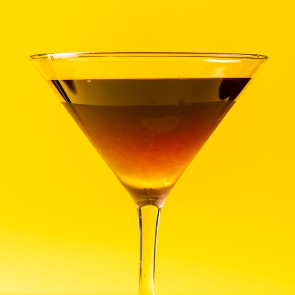 Martini glass filled with a dark amber drink, set against a vibrant yellow background.