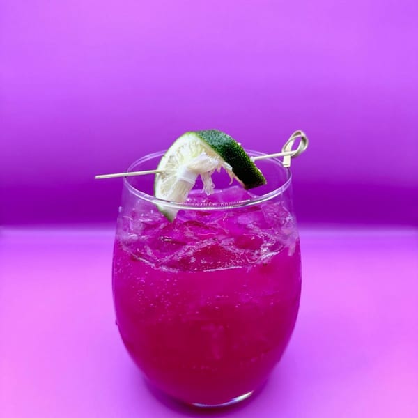 A vibrant pink cocktail with ice and a lime wedge garnish, set against a purple background.