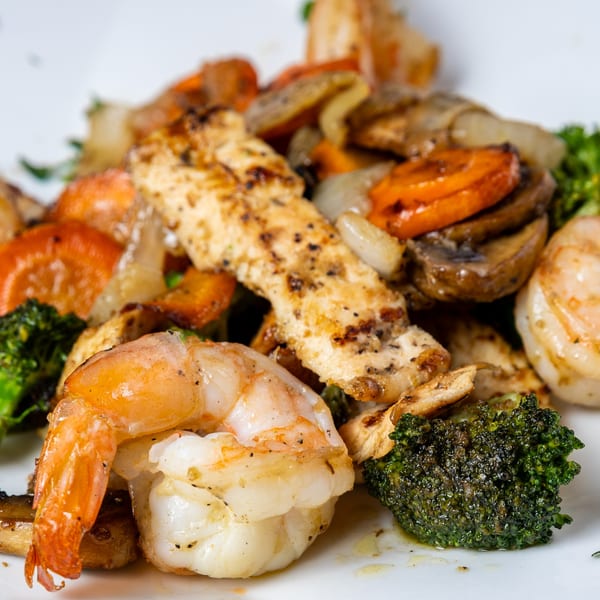 Close-up of grilled chicken, shrimp, broccoli, carrots, onions, and mushrooms on a white plate.