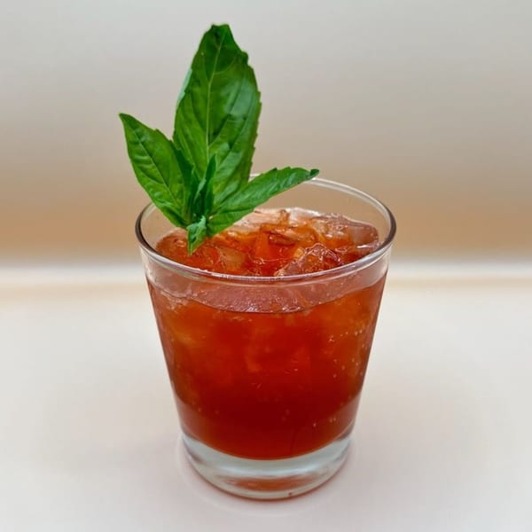 A vibrant red iced drink in a clear glass, garnished with fresh green basil leaves.